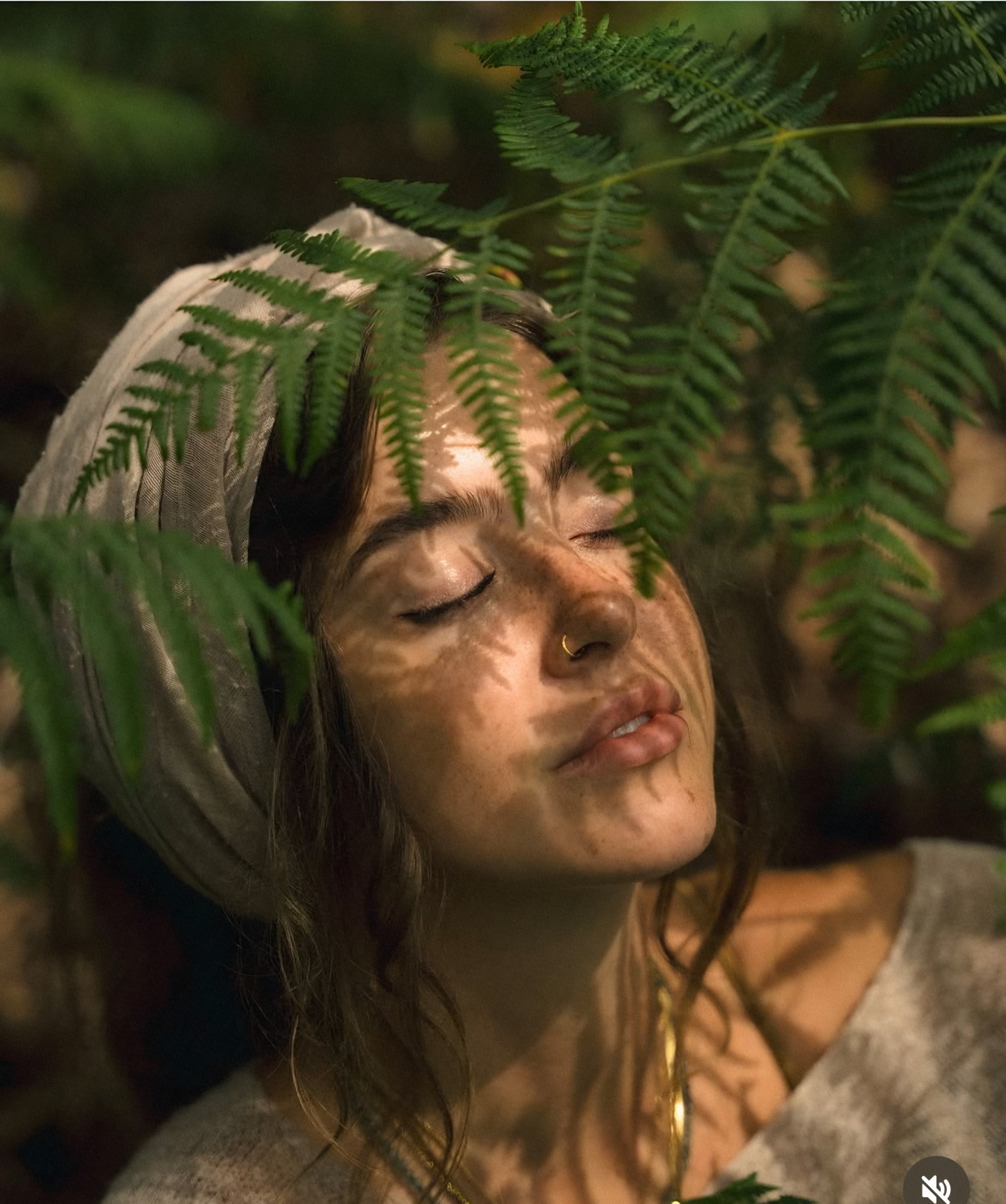 A woman with eyes closed beneath fern fronds, dappled sunlight on her face