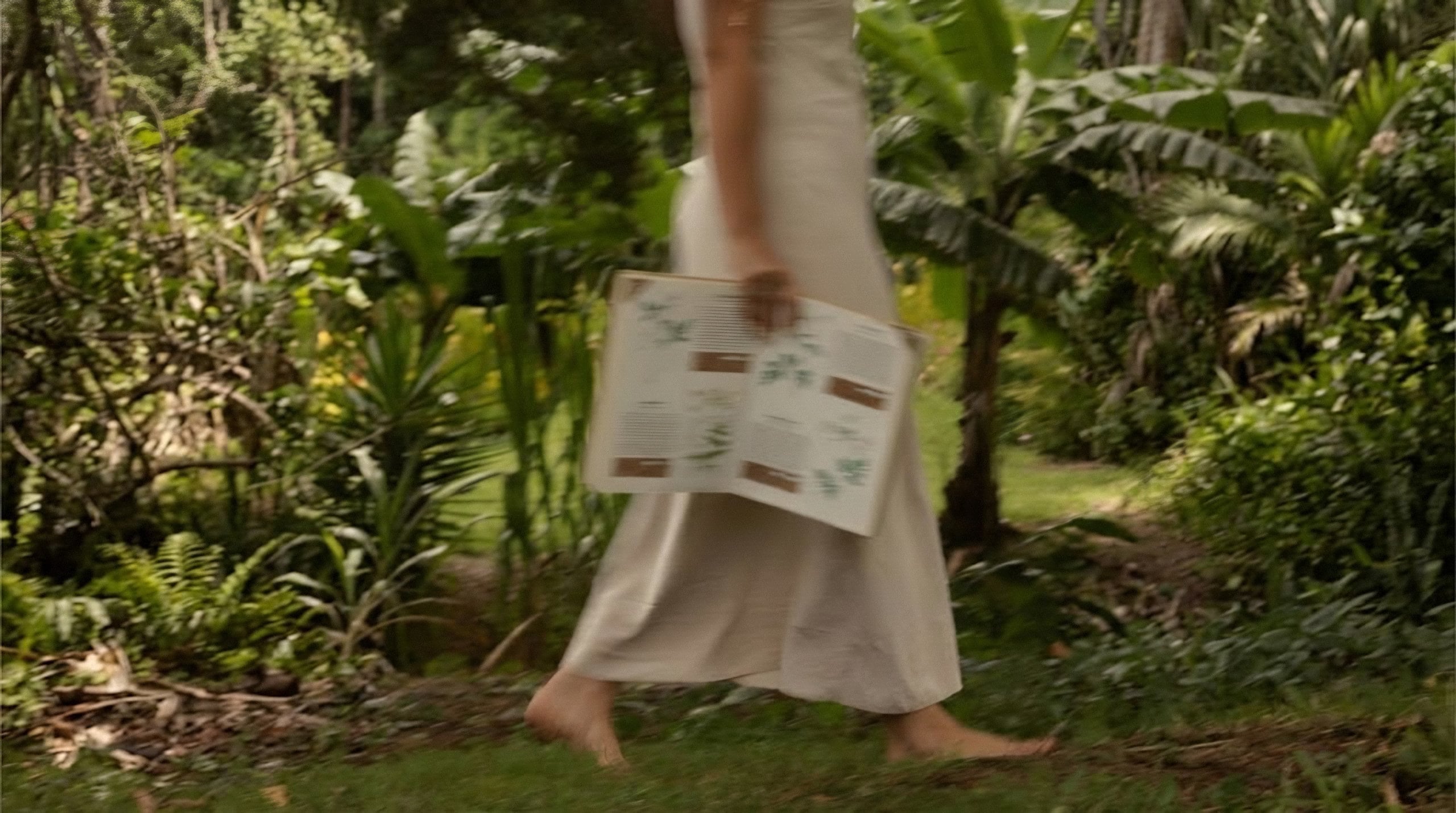 A woman walks barefoot through a garden holding an open botanical book